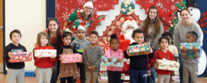 College students stand with preschoolers holding gift boxes next to Santa with a backdrop of festive, holiday decorations.