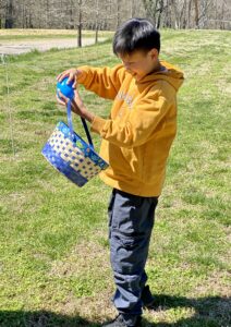 Young boy wearing yellow sweatshirt and holding a multi-color basket holds a large, blue plastic egg.