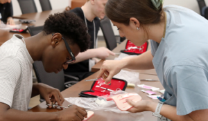 A high school student practices stitching sutures with a surgical resident 