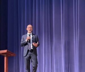 President Paino in blue suit with microphone on stage in GW Hall