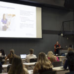 students in a classroom look at a presentation at the front of the room where there's also a speaker
