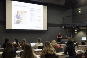 students in a classroom look at a presentation at the front of the room where there's also a speaker