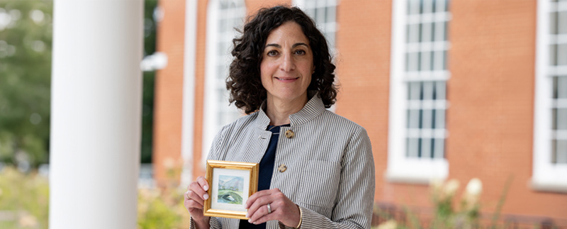 woman standing in front of brick building holds a small painting in a frame