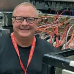 A man with glasses and dark top stands in front of machines with many wires