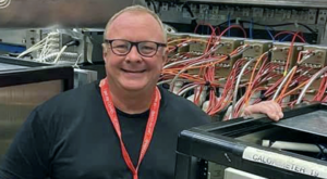 A man with glasses and dark top stands in front of machines with many wires