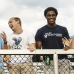 male and female students with University of Mary Washington t-shirts laugh while standing at the railing of an outdoor sports stadium