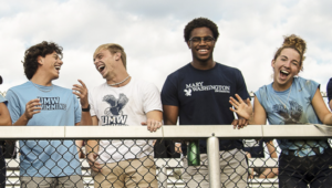 male and female students with University of Mary Washington t-shirts laugh while standing at the railing of an outdoor sports stadium