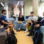 four students and a professor sit in colorful chairs with laptops inside the Hurley Convergence Center on UMW campus