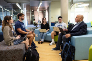 four students and a professor sit in colorful chairs with laptops inside the Hurley Convergence Center on UMW campus