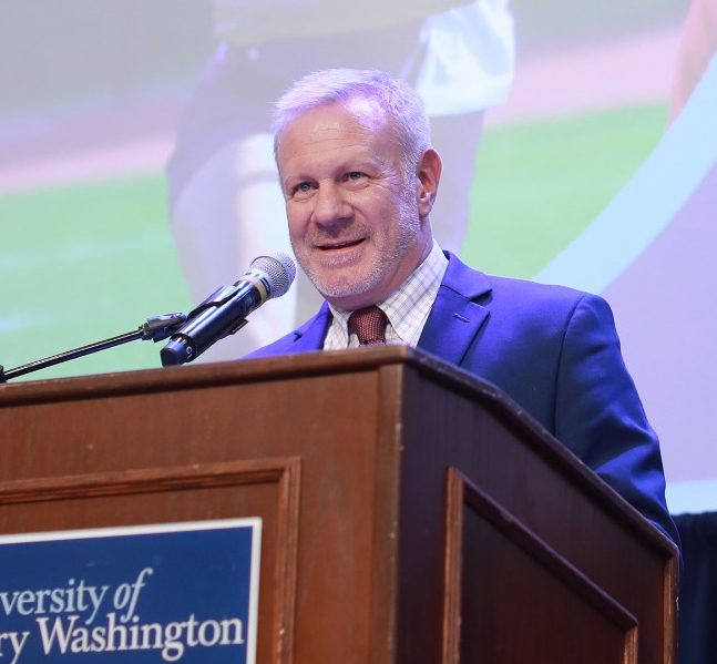 man with jacket and tie speaks into a microphone behind a podium