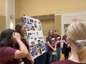 several students stand near a poster on an easel in a ballroom