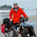 man stands next to a bicycle with a red jacket on the sand at the ocean